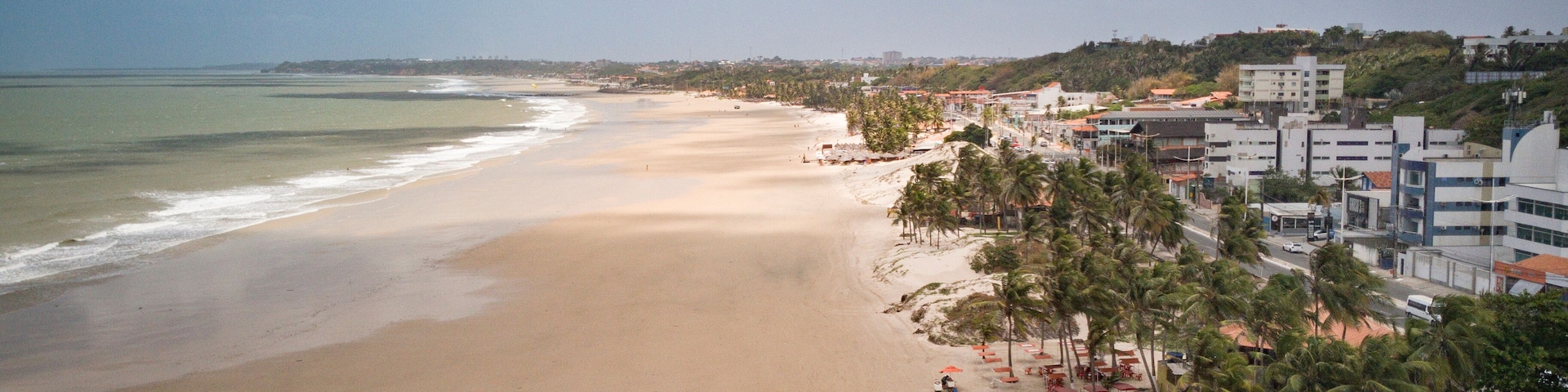 Una amplia playa de arena con olas suaves, bordeada por palmeras y edificios frente al mar.