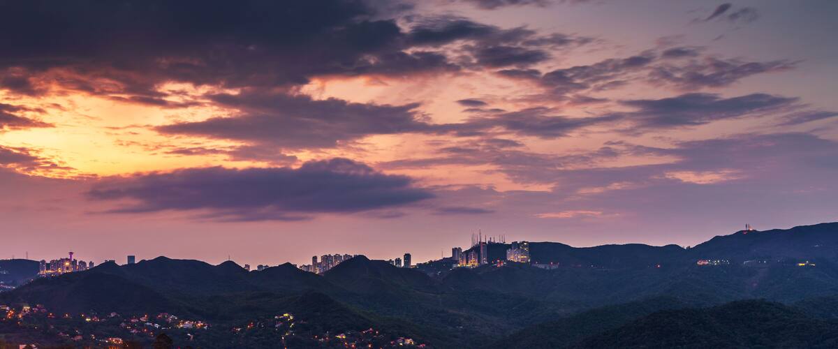 A city skyline at dusk with illuminated buildings against a backdrop of hills and a colorful sky.