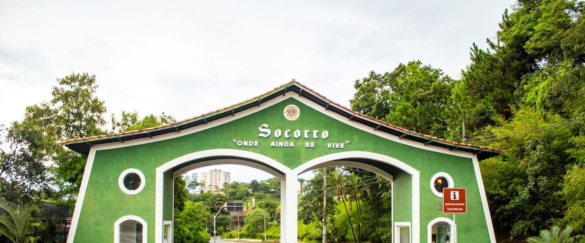 A green archway with 'Socorro' and 'Onde Ainda Se Vive' text, a road, and lush greenery.
