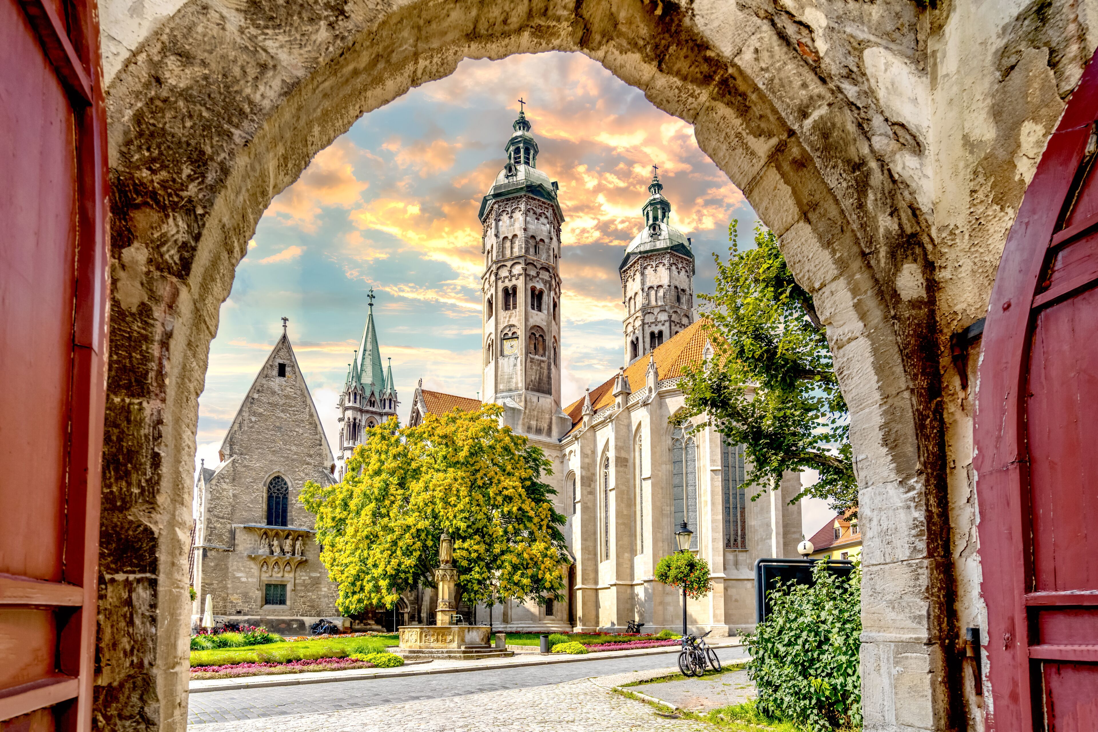 An archway framing a historic church with twin towers and a large tree in the foreground.
