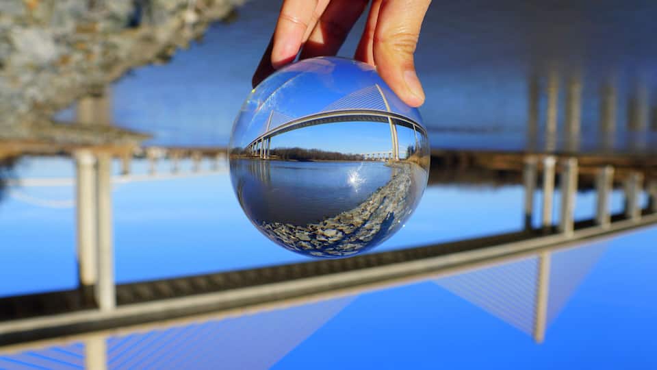 A hand holding a clear sphere reflecting a bridge and a body of water.