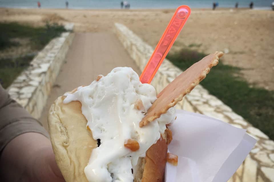 A person holding a sandwich with a white sauce and a piece of meat, on a beach with a path and ocean in the background.
