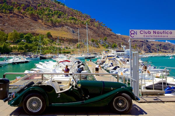Un'auto verde d'epoca parcheggiata in una marina, con barche e un cartello per Club Nautico Castellammare.