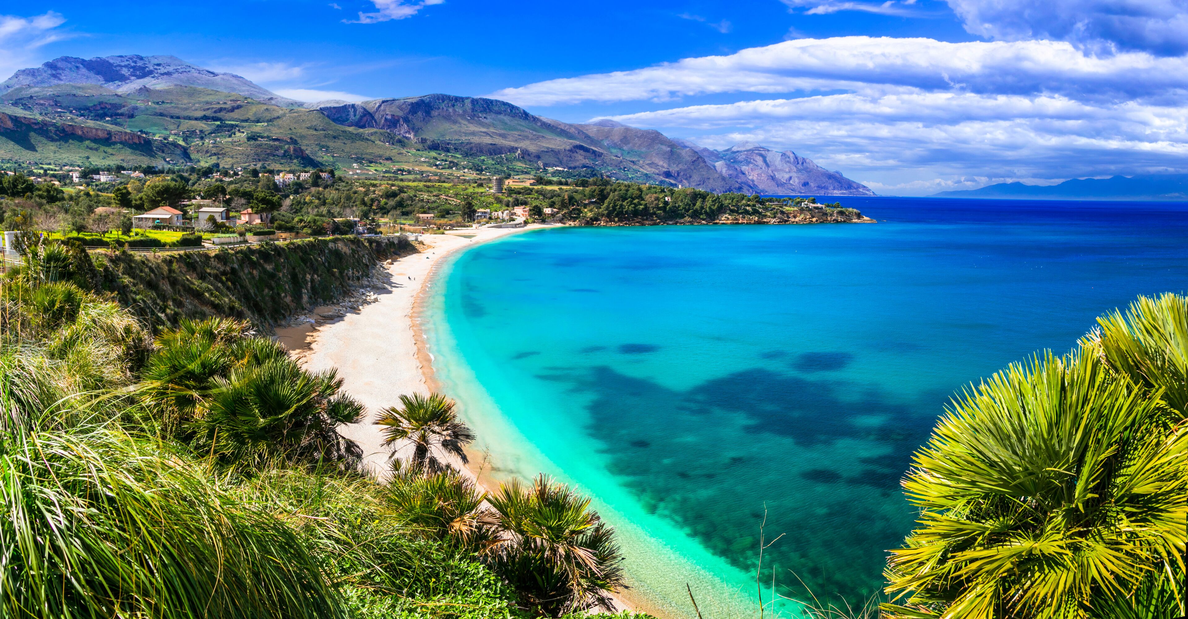 A coastal landscape with a turquoise beach, green hills, and a mountain range.