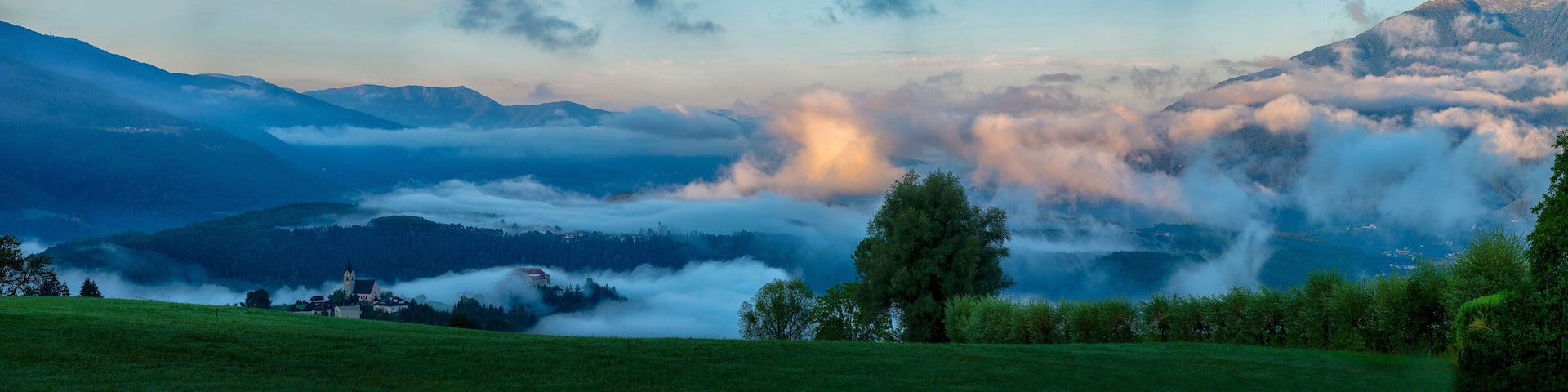 A mountainous landscape with a church, trees, and a valley shrouded in clouds.