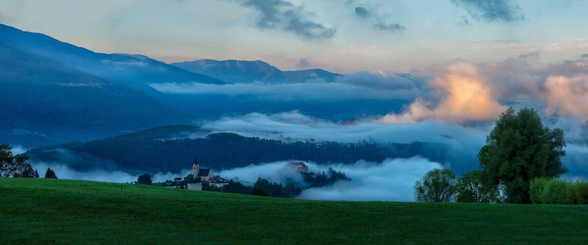 A mountainous landscape with a church, trees, and a valley shrouded in clouds.