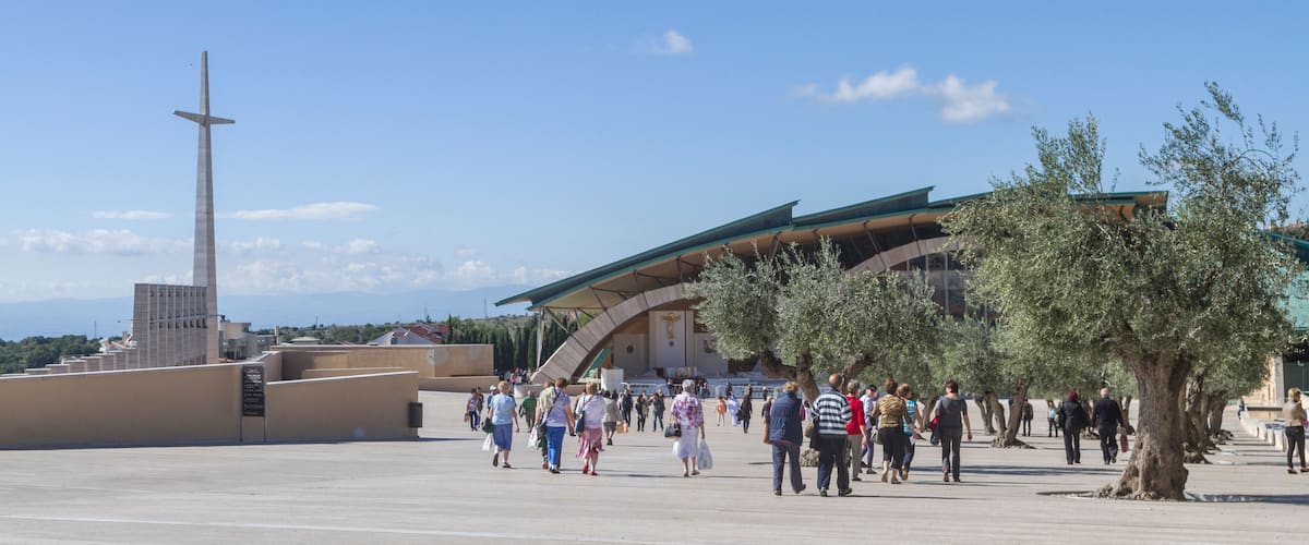 A large open plaza with a tall monument, a modern building with a green roof, and olive trees.