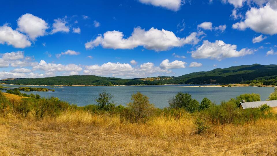 A lake surrounded by hills and dry grassland with a few buildings in the distance.