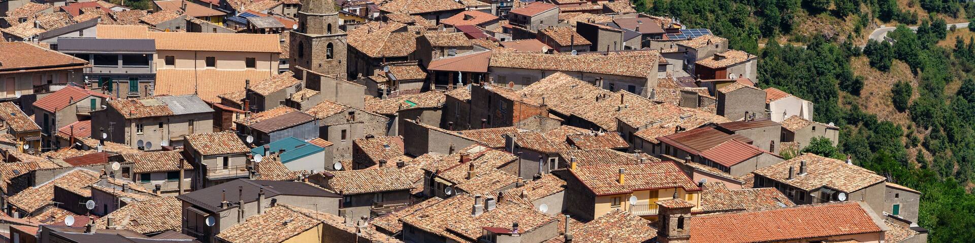 A densely packed hillside village with terracotta-roofed buildings, a prominent church tower, and lush green hills in the background.