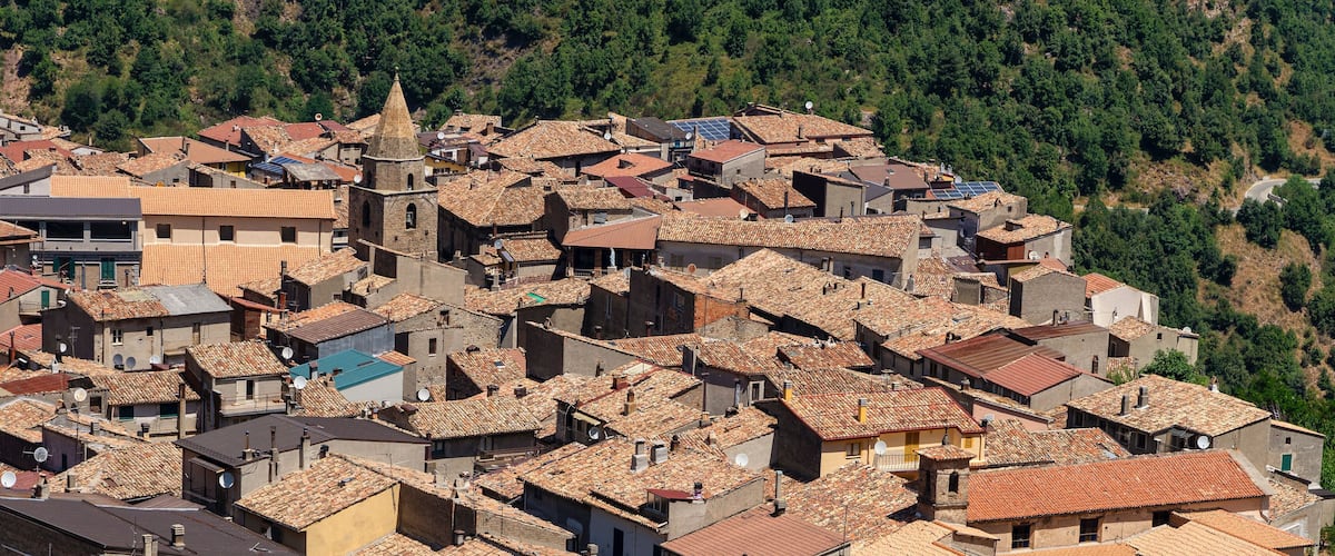 A densely packed hillside village with terracotta-roofed buildings, a prominent church tower, and lush green hills in the background.