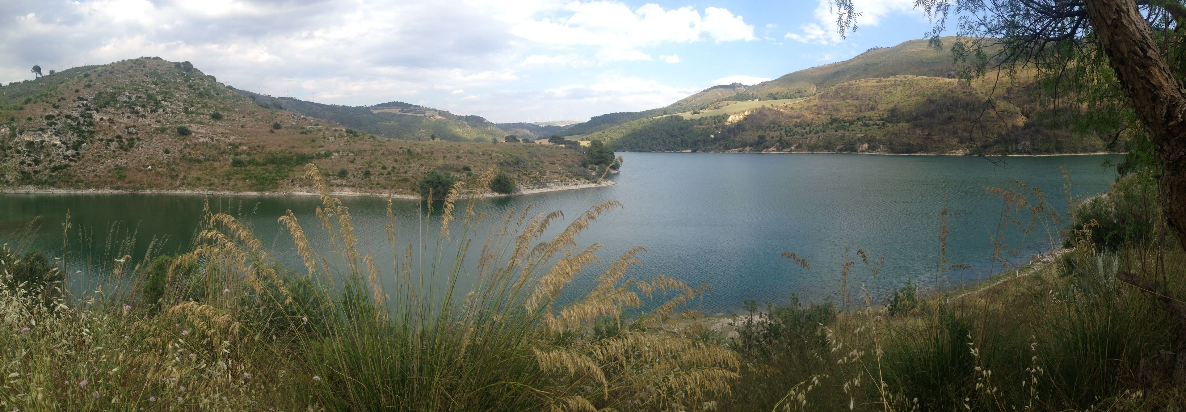 A lake surrounded by hills with wind turbines in the distance.