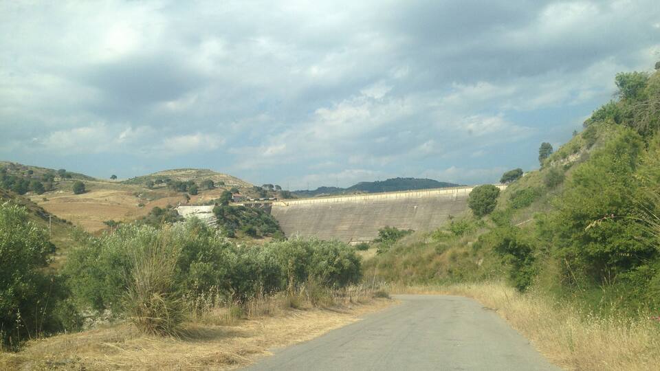 A rural road flanked by dry grass and shrubs, leading towards a large dam with a reservoir.