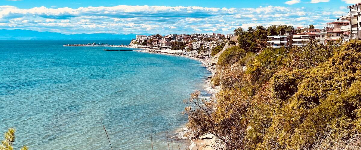 Coastal view of a Mediterranean seaside town with turquoise water, lush greenery, and clear blue sky. Residential buildings line the coastline, creating a picturesque summer atmosphere in a Greek reso