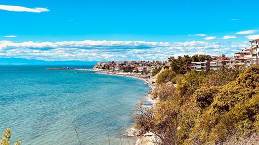 Coastal view of a Mediterranean seaside town with turquoise water, lush greenery, and clear blue sky. Residential buildings line the coastline, creating a picturesque summer atmosphere in a Greek reso