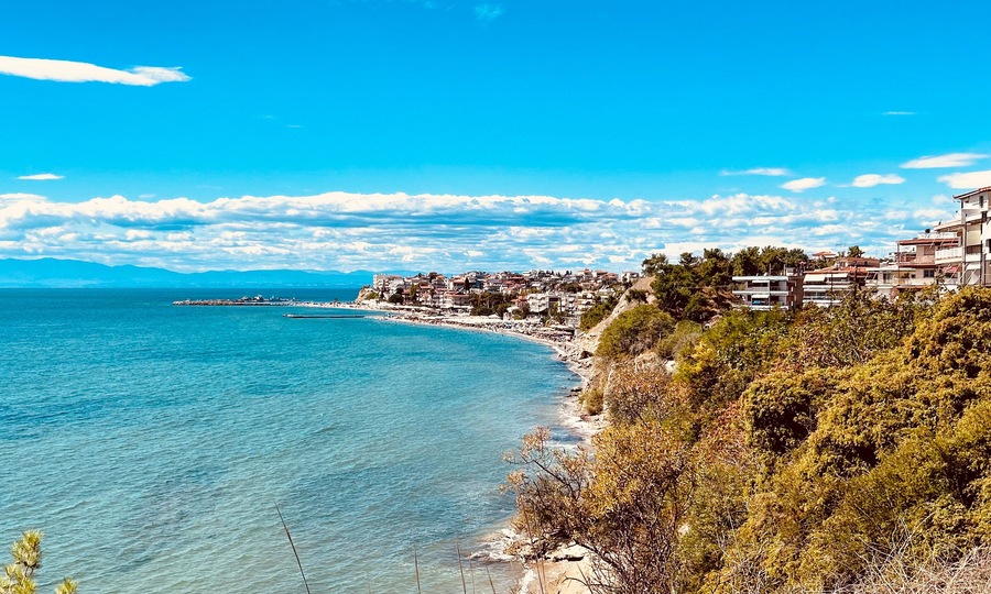 Coastal view of a Mediterranean seaside town with turquoise water, lush greenery, and clear blue sky. Residential buildings line the coastline, creating a picturesque summer atmosphere in a Greek reso