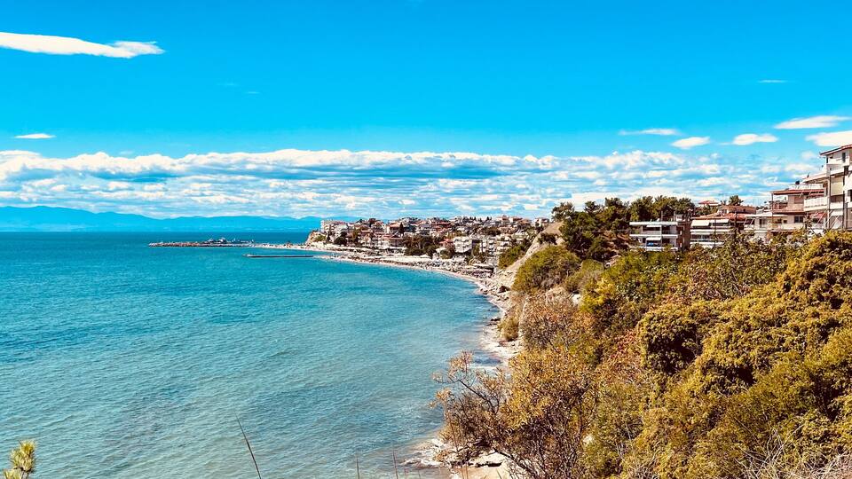 Coastal view of a Mediterranean seaside town with turquoise water, lush greenery, and clear blue sky. Residential buildings line the coastline, creating a picturesque summer atmosphere in a Greek reso