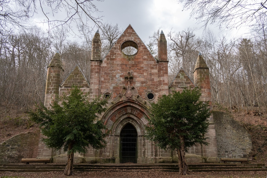 A historic stone church with a pointed arch entrance, flanked by two trees, surrounded by a forested area.