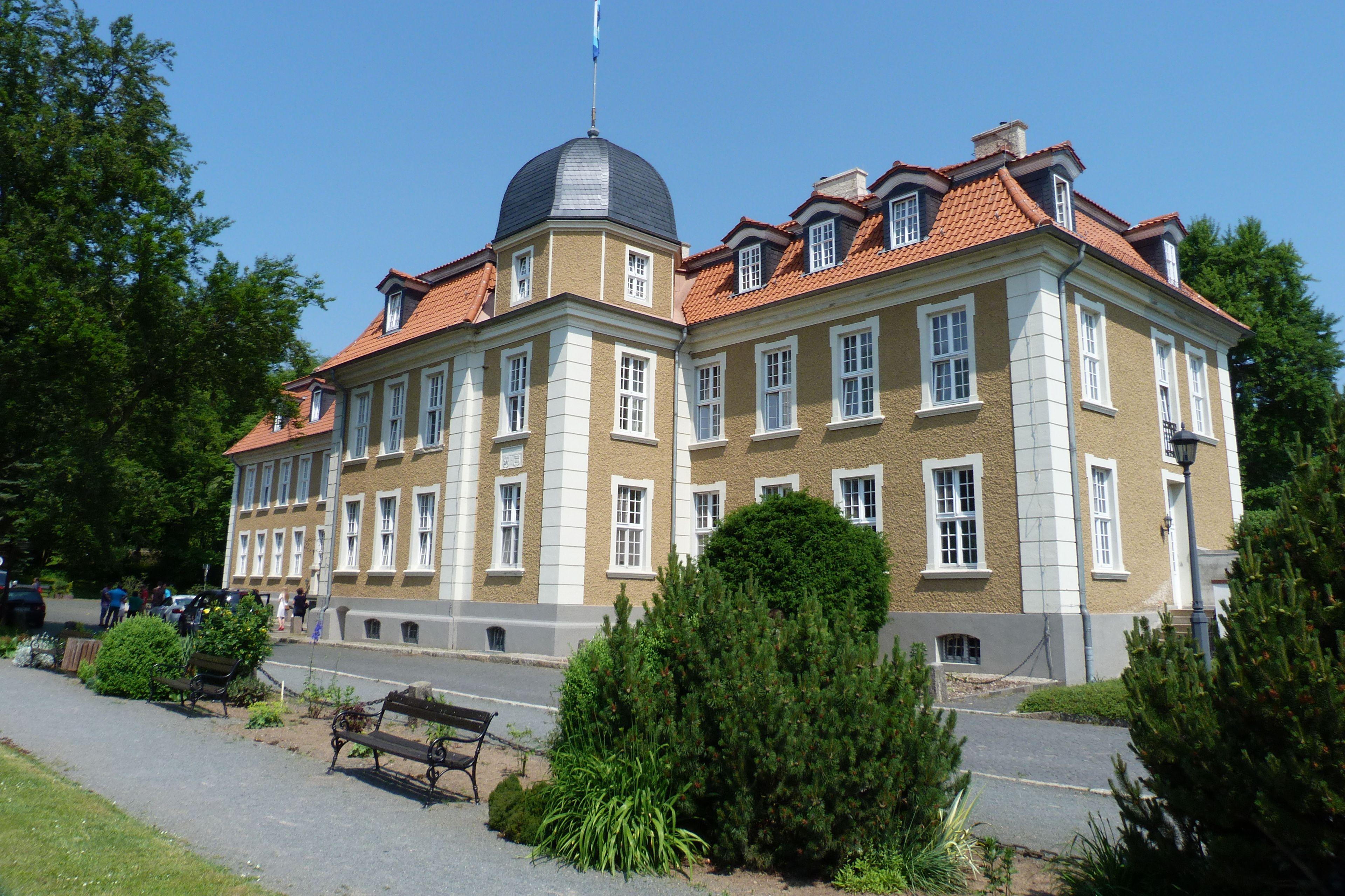 A large, historic building with a dome and red-tiled roof, surrounded by greenery and a paved walkway.
