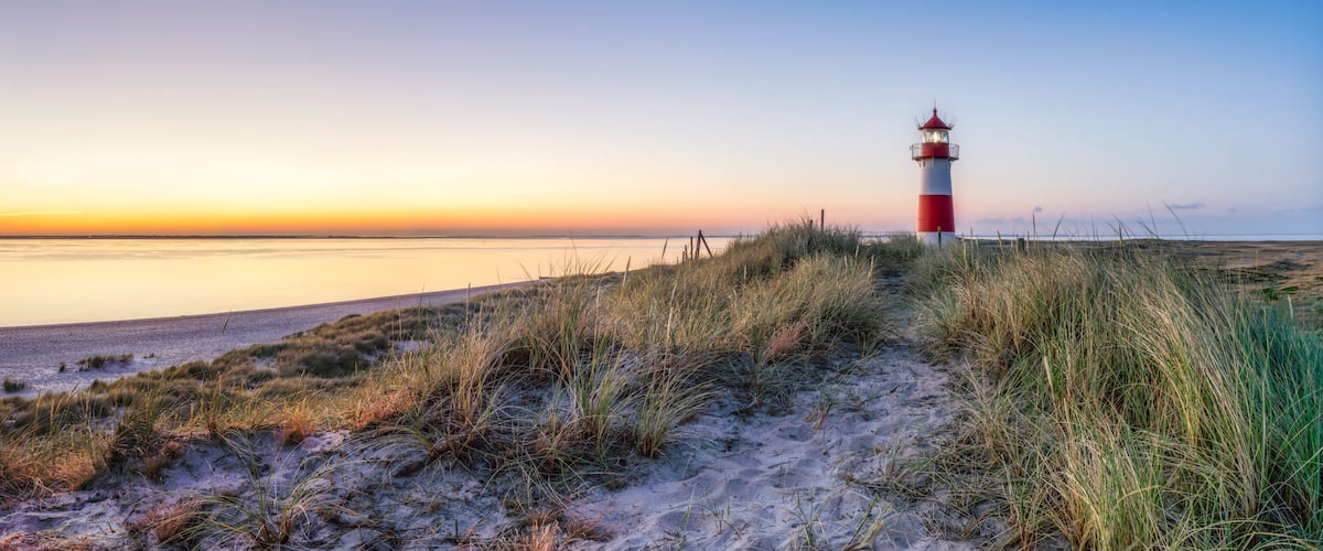 A lighthouse on a sandy beach with grass in the foreground and a calm sea in the background.