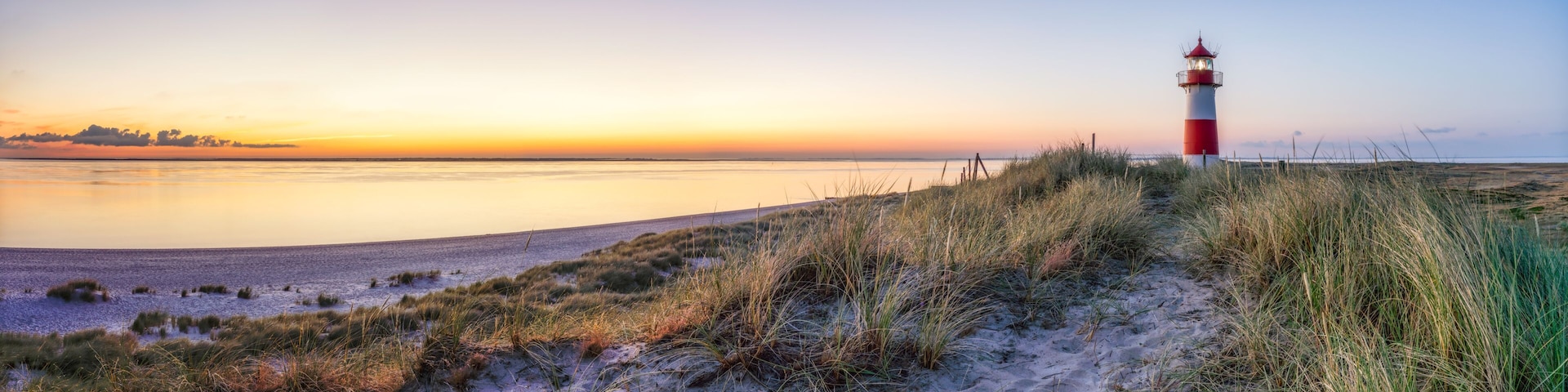 A lighthouse on a sandy beach with grass in the foreground and a calm sea in the background.