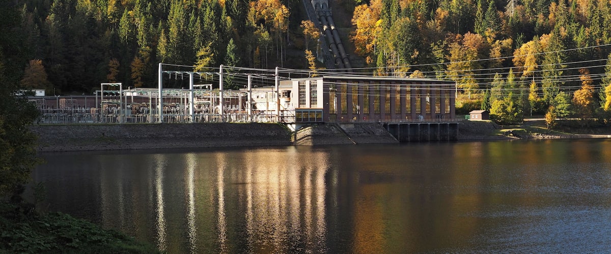 A hydroelectric dam with a water reservoir, surrounded by autumn-colored trees and a forested hillside.