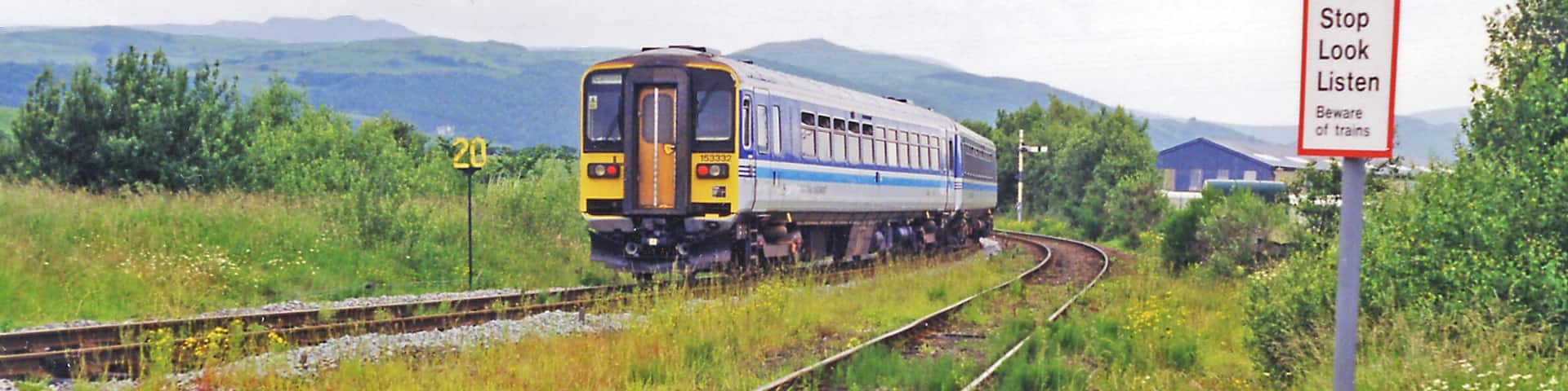 Foxfield: train leaving for Whitehaven, 1998. View northwards, up Dunnerdale towards Coniston ahead - to which the now vanished ex-Furness Railway branch used to go until 6/4/62, the passenger service having ceased since 6/10/58. The Class 153 DMU, on a Carnforth - Barrow-in-Furness - Whitehaven service on the ex-Furness Railway main line, will very soon curve left to cross the Duddon Viaduct, then go south to Millom before continuing up the coast to Whitehaven. Note the weed-strewn tracks. In the distance are Thwaites Fell (1,001 ft.), Burn Moor (1,780 ft.) and Whitwell (1,861 ft.).