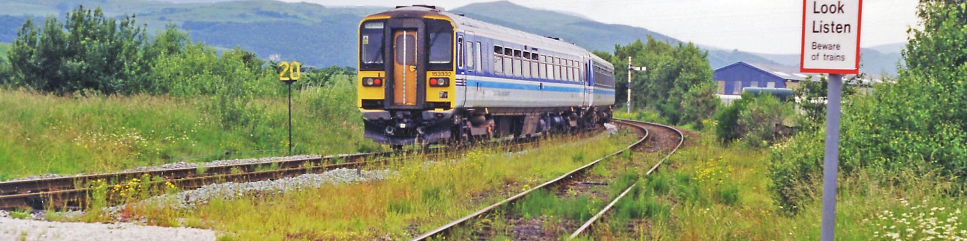 Foxfield: train leaving for Whitehaven, 1998. View northwards, up Dunnerdale towards Coniston ahead - to which the now vanished ex-Furness Railway branch used to go until 6/4/62, the passenger service having ceased since 6/10/58. The Class 153 DMU, on a Carnforth - Barrow-in-Furness - Whitehaven service on the ex-Furness Railway main line, will very soon curve left to cross the Duddon Viaduct, then go south to Millom before continuing up the coast to Whitehaven. Note the weed-strewn tracks. In the distance are Thwaites Fell (1,001 ft.), Burn Moor (1,780 ft.) and Whitwell (1,861 ft.).