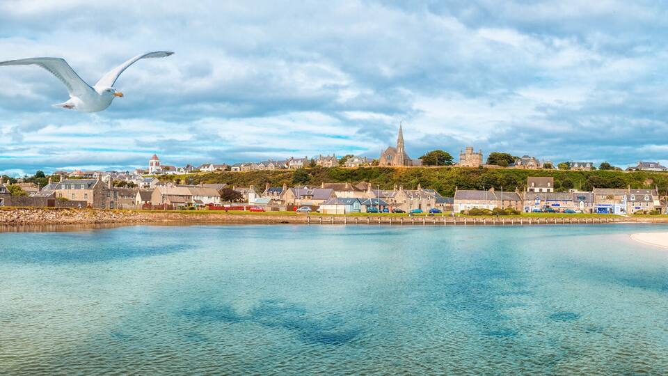 A coastal town with a river, a sandy beach, and a hill with buildings and a church.