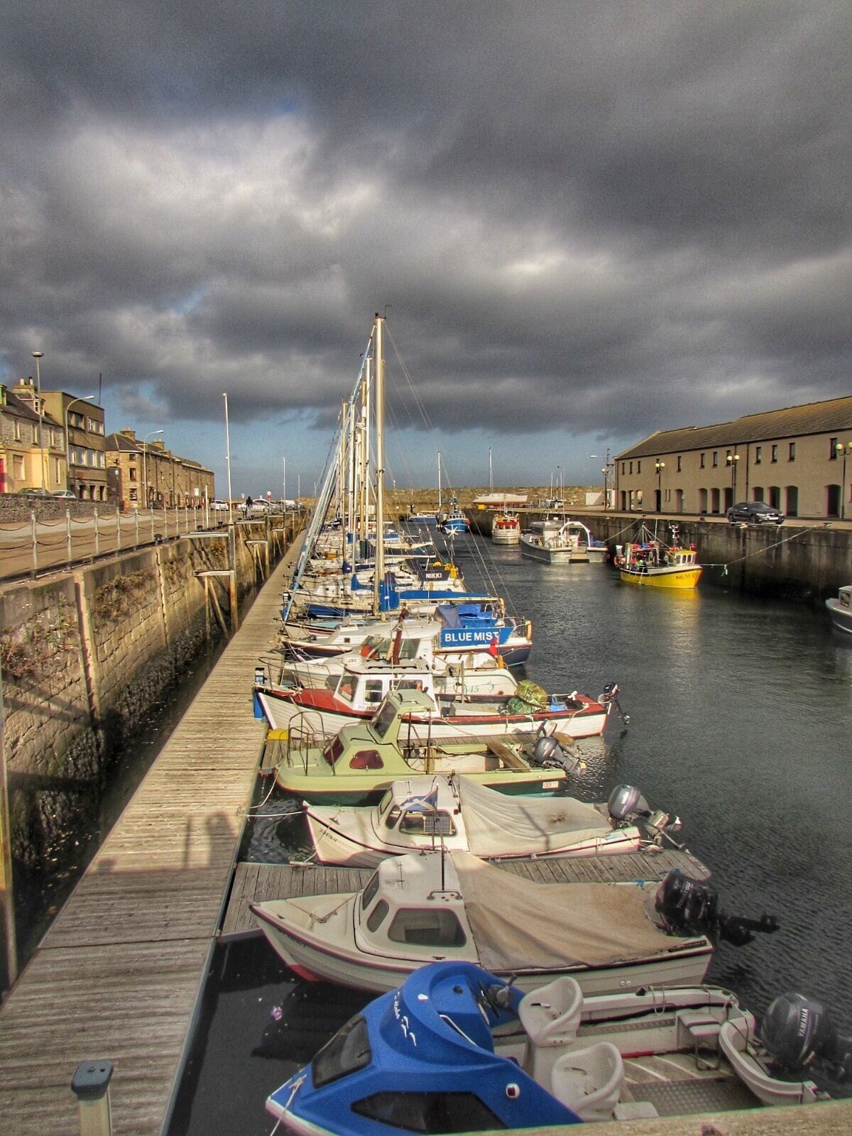 A harbor with various boats docked along a wooden pier, under a cloudy sky.