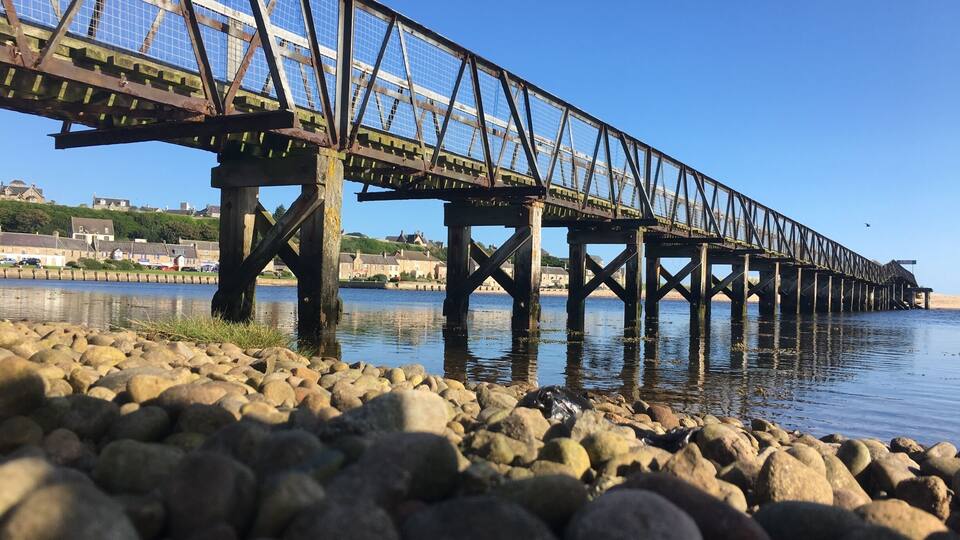 A steel bridge over a calm river with a rocky shoreline.