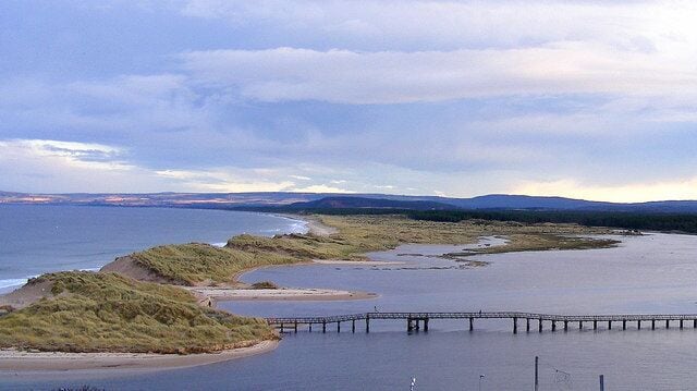 A coastal landscape with a bridge over water, grassy dunes, and distant hills.
