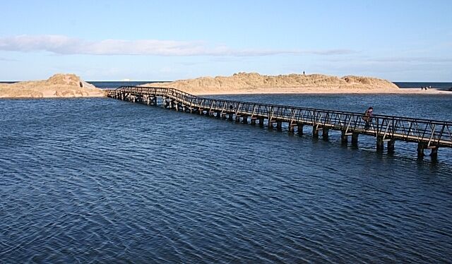 A long wooden pier extending into calm water, with sandy dunes in the background.