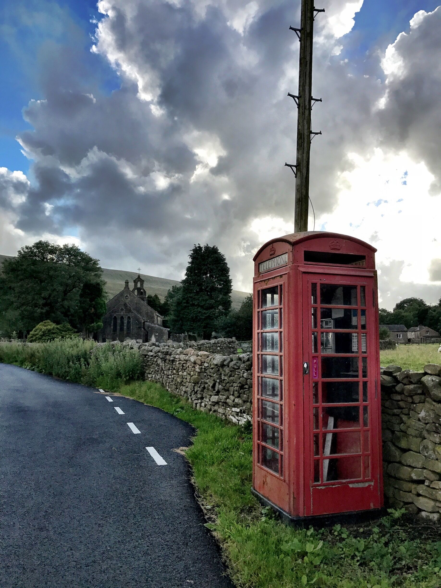 A red telephone booth on a rural road with a stone wall and a church in the background.