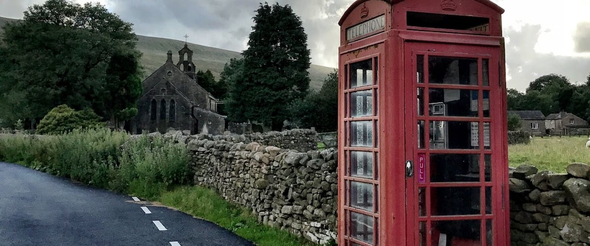 A red telephone booth on a rural road with a stone wall and a church in the background.