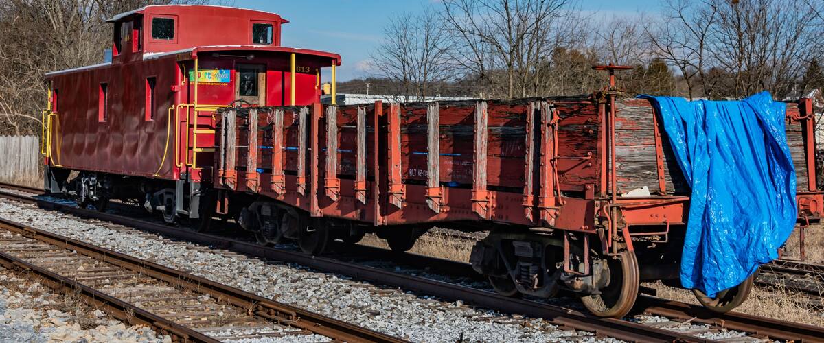 A red train car with a blue tarp on the right side, parked on a railway track with trees in the background.