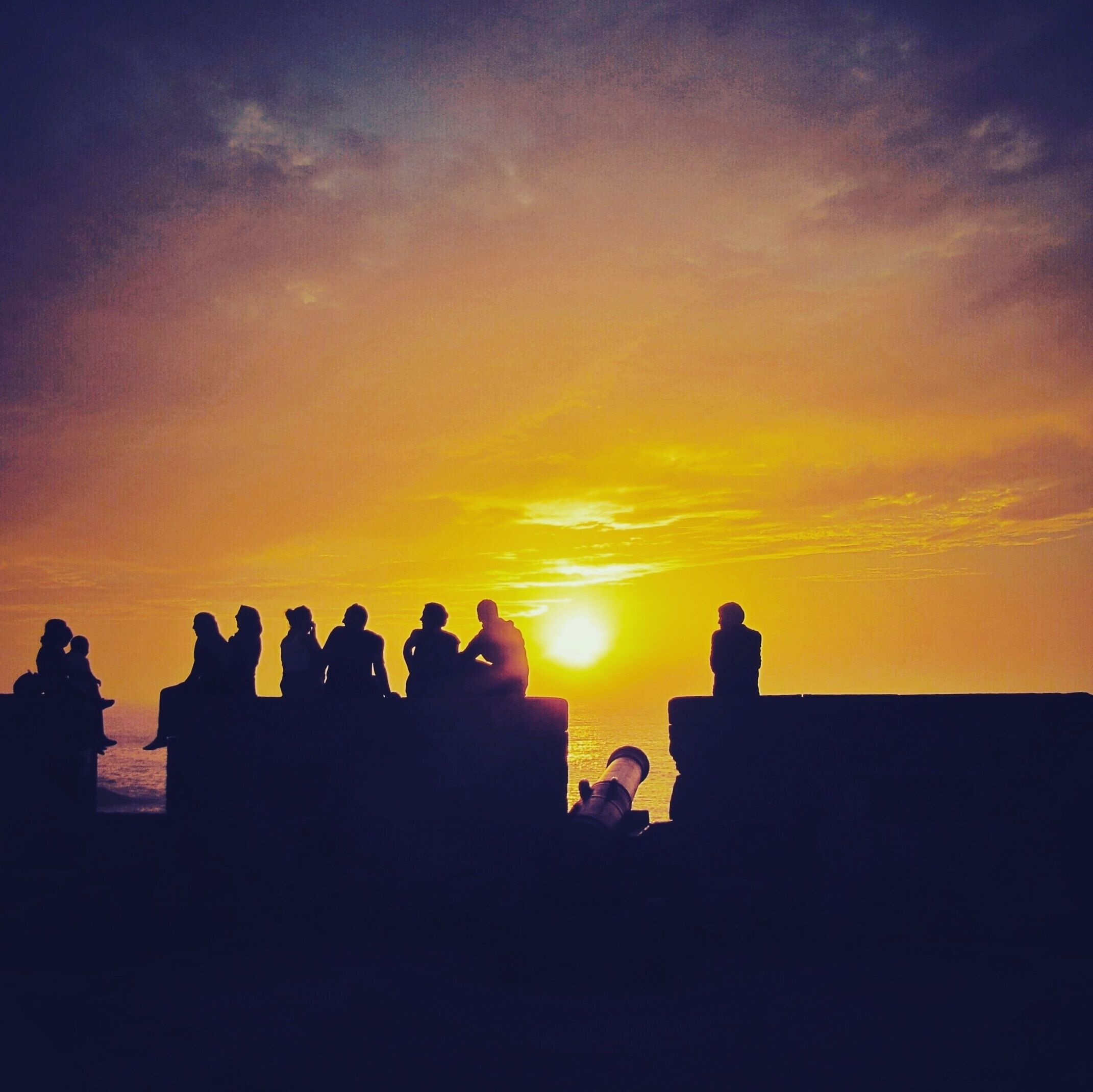 Siluetas de personas sentadas en un saliente con vista a una puesta de sol sobre el océano.