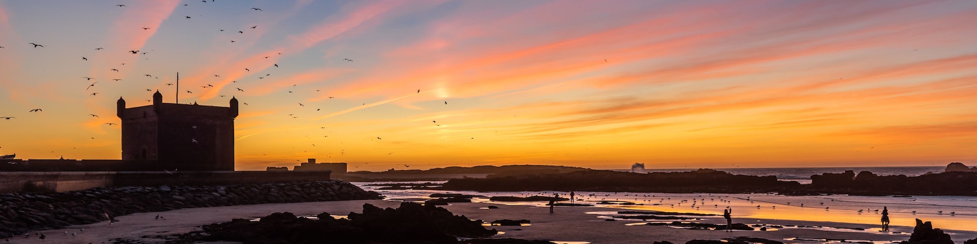 A sunset over a rocky beach with birds flying, a tower, and people walking.