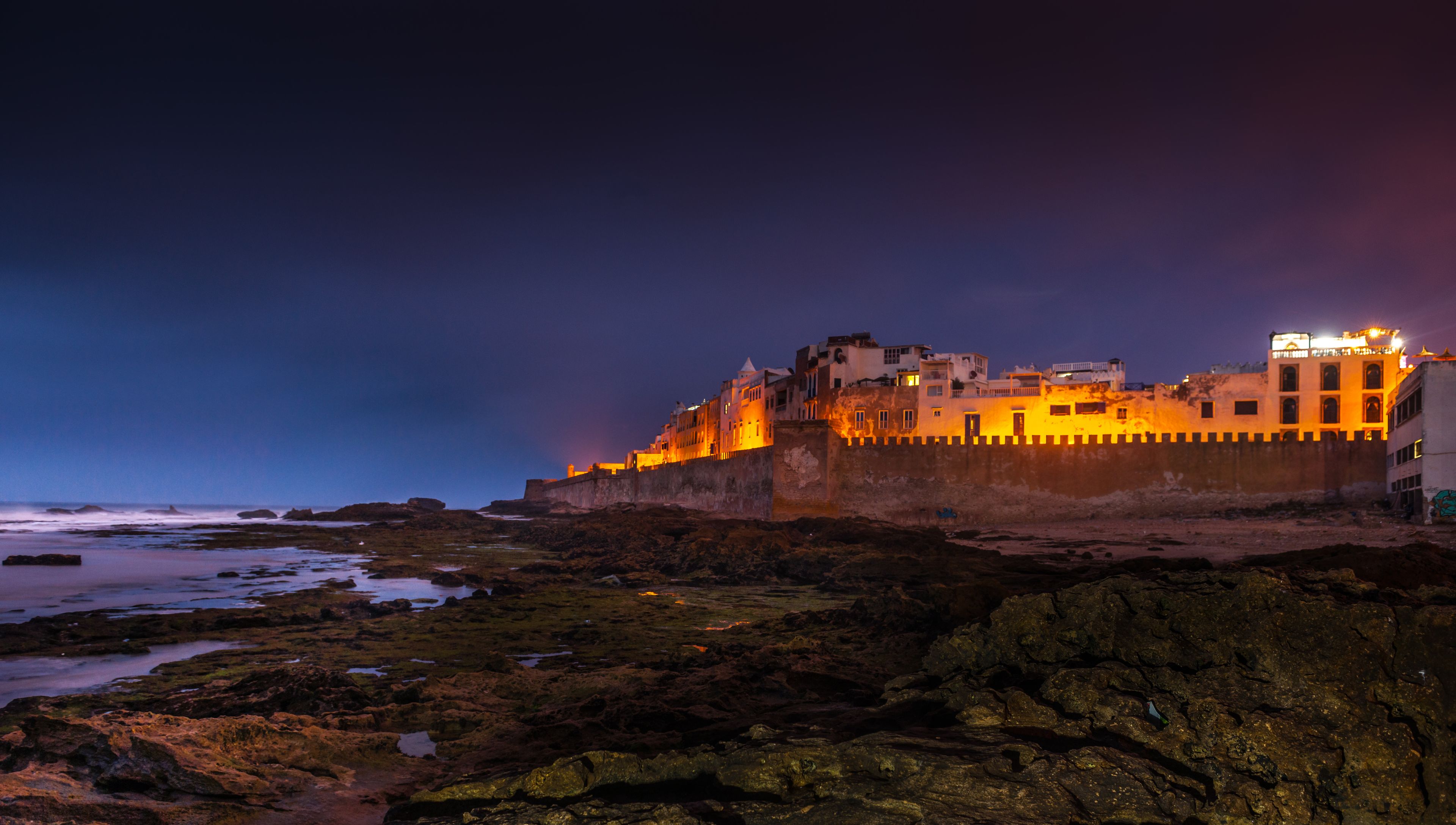 A coastal fortress with illuminated walls and a rocky shoreline at dusk.