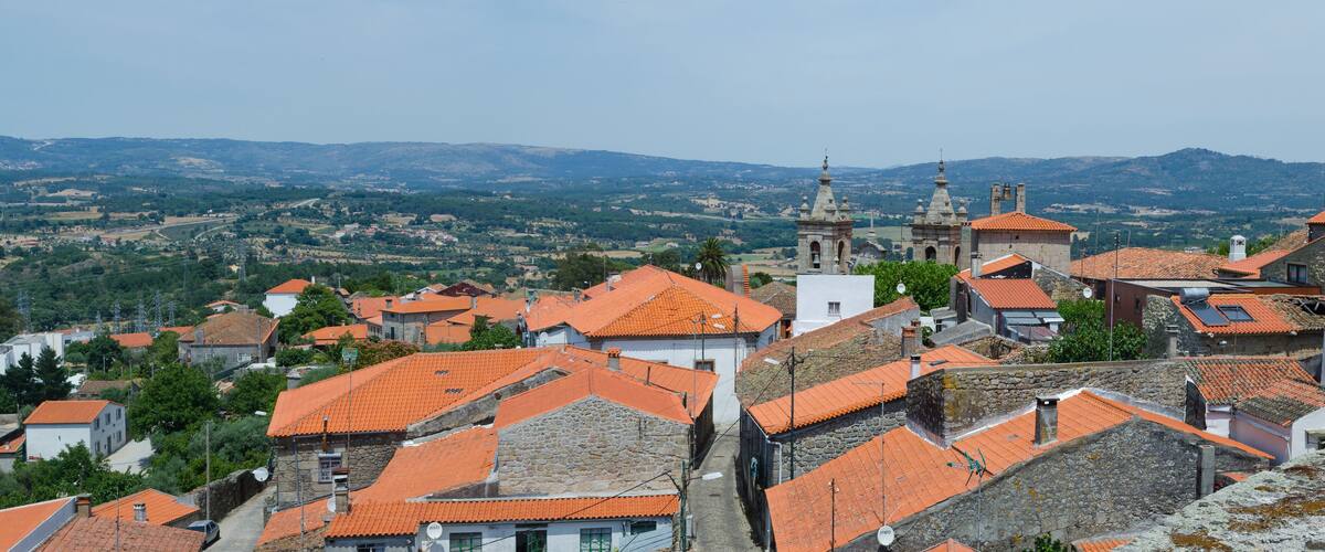 A town with orange-roofed buildings, a prominent church with two towers, and a mountainous backdrop.