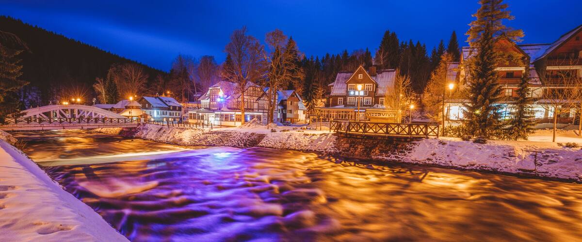 A snow-covered riverbank with a wooden bridge and illuminated buildings in the background.