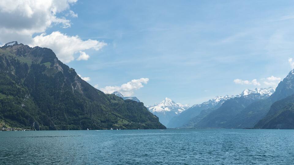 A calm lake surrounded by steep, forested mountains with snow-capped peaks in the distance.