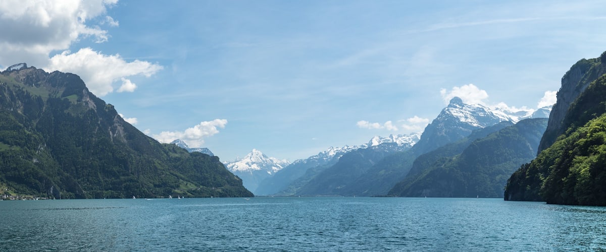 A calm lake surrounded by steep, forested mountains with snow-capped peaks in the distance.