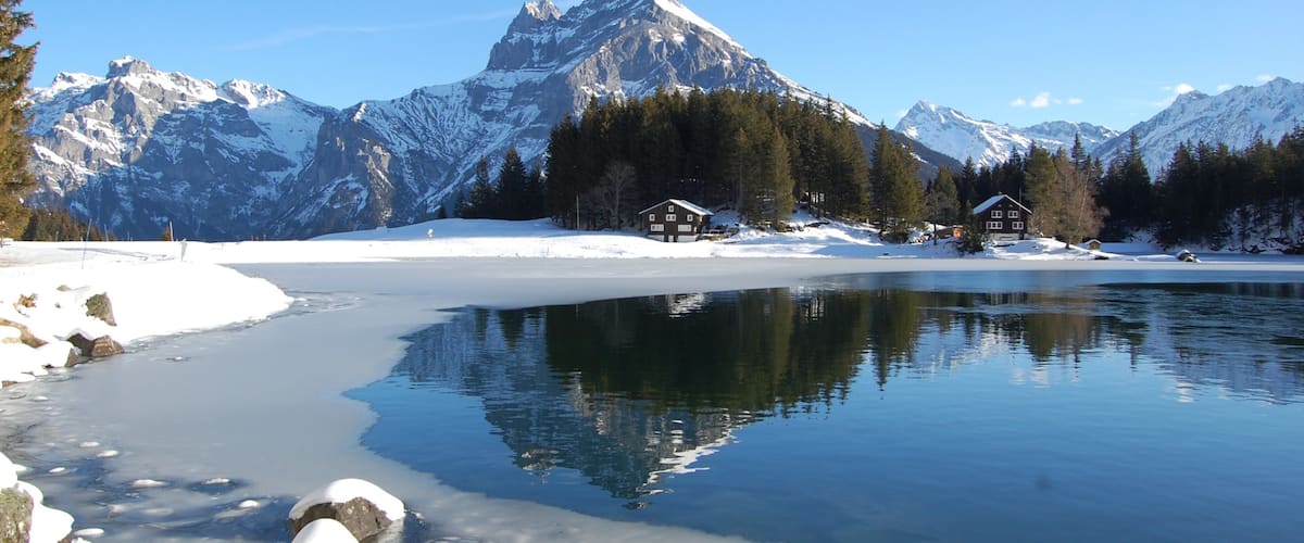 A frozen lake with snow-covered mountains in the background and two wooden cabins near the shore.