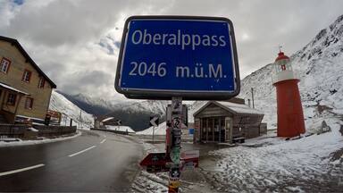 A sign reading "Oberalppass 2046 m.U.M." with a red lighthouse and snow-covered mountains in the background.