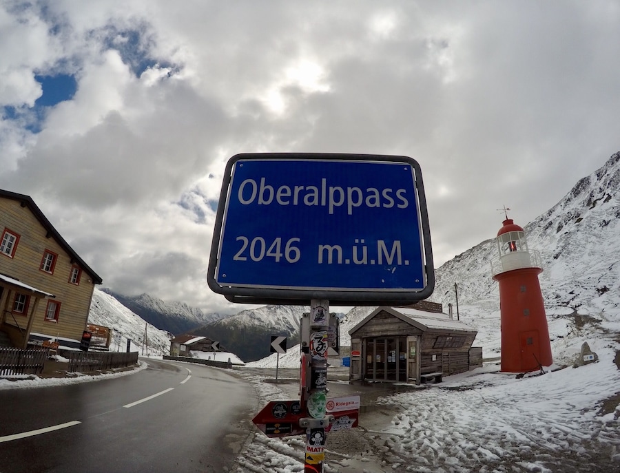 A sign reading "Oberalppass 2046 m.U.M." with a red lighthouse and snow-covered mountains in the background.