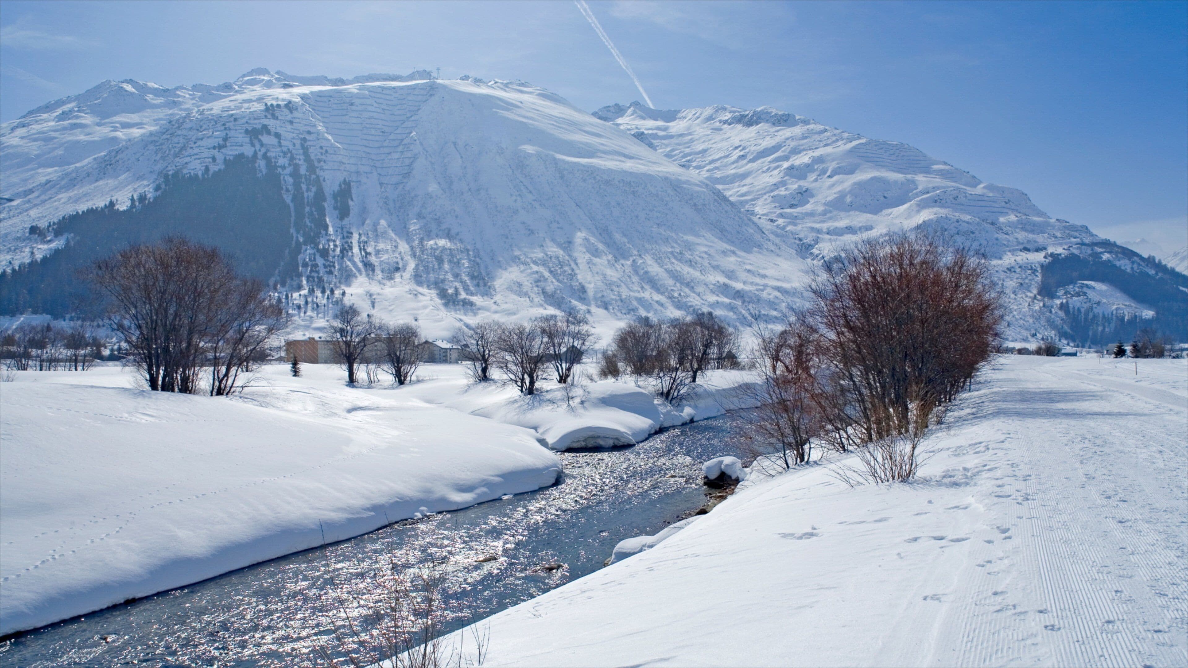 A snow-covered landscape with a river, trees, and mountains in the background.