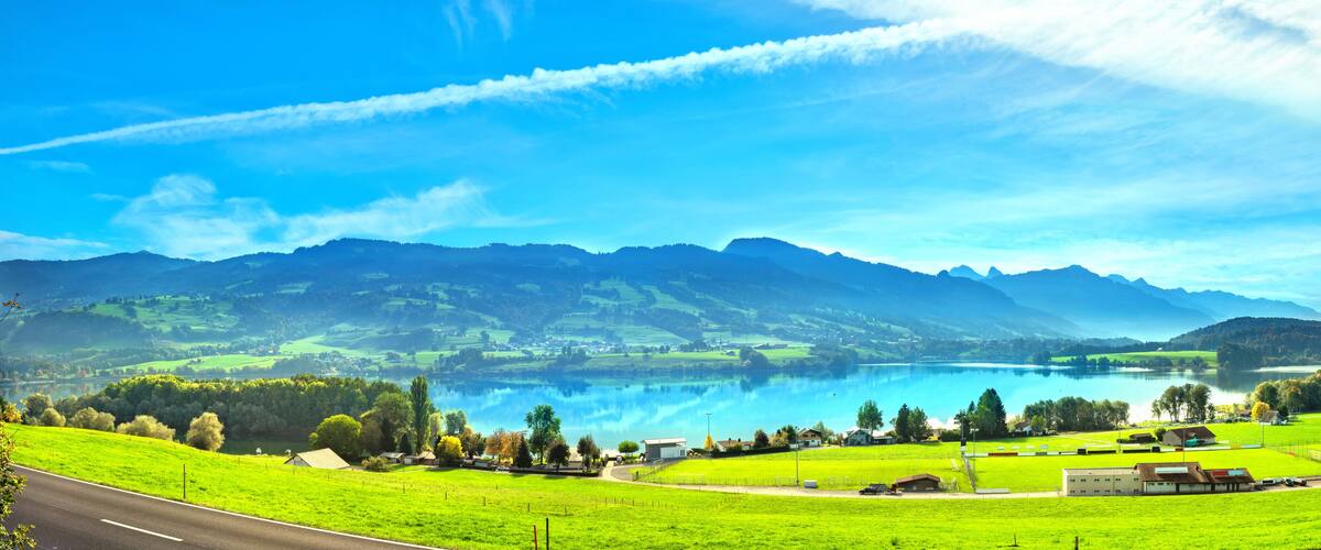A lake surrounded by mountains, a road, and green fields.