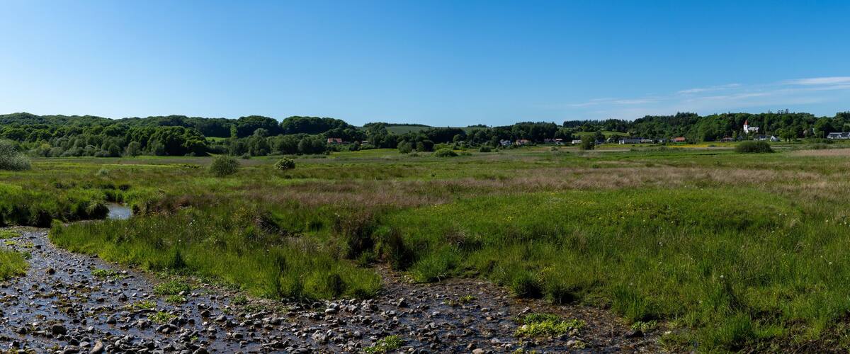 A rural landscape with a river, grassy fields, and a small village in the distance.