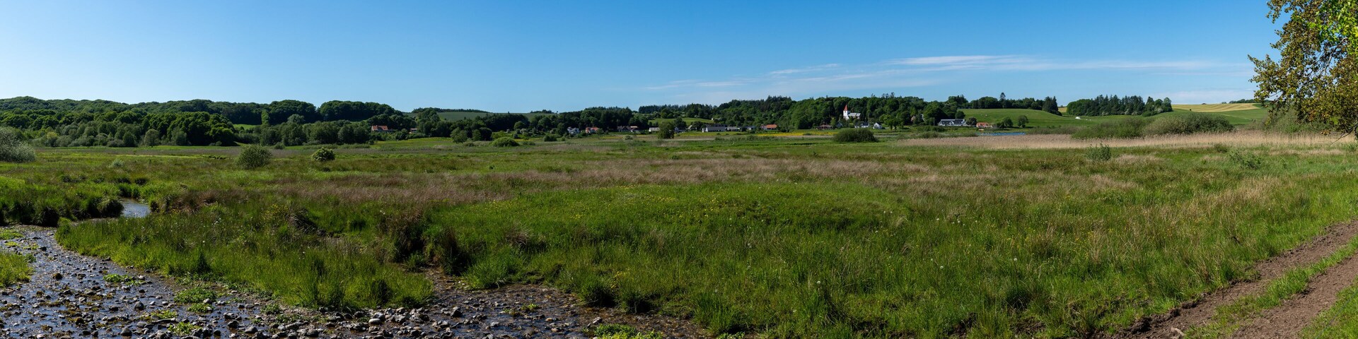 A rural landscape with a river, grassy fields, and a small village in the distance.