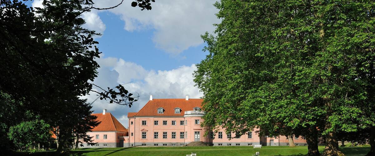 A large pink building with a red roof, surrounded by trees and a pond reflecting the structure.